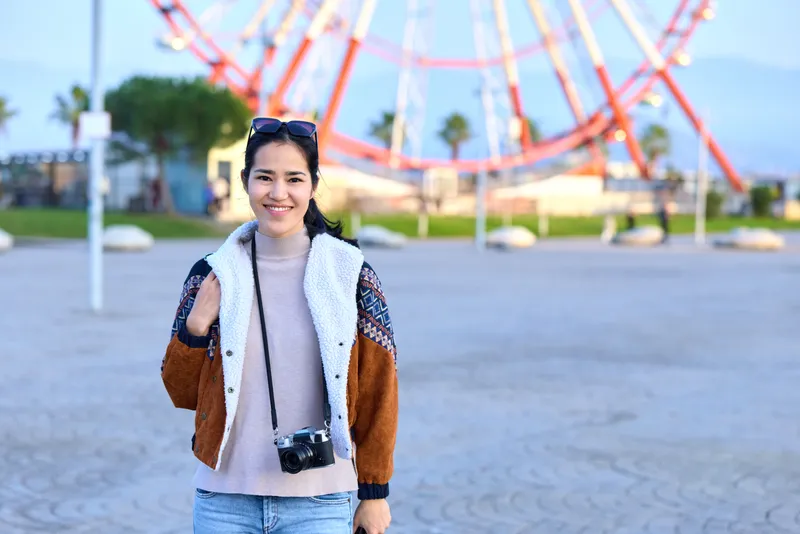 Smiling woman in a colorblock jacket and cozy sweater with a camera hanging, standing outdoors near a ferris wheel in a sunny park, capturing a casual travel moment and stylish everyday vibe.
