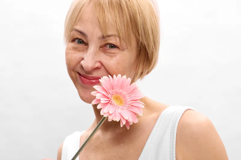Elderly woman with short blonde hair has a gentle smile, holding a pink flower blossom near her face against a clean white background, beauty portrait closeup
