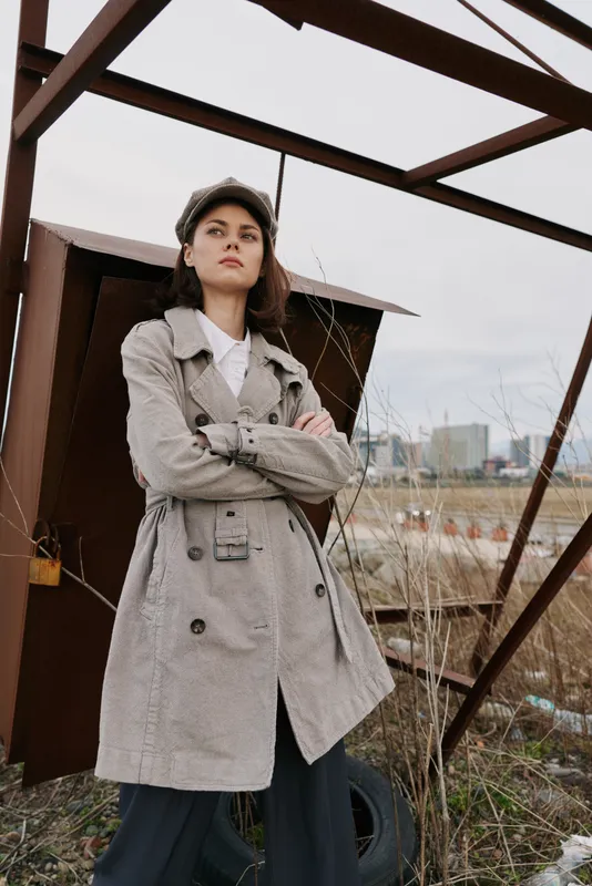 Confident young woman in a plaid trench coat and matching cap stands outdoors near metal structure with urban background. Fashion concept with casual autumn outfit and serious mood.