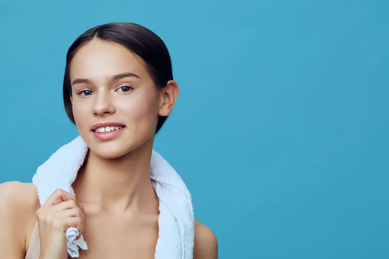 Young woman with fresh skin and a towel on her shoulders, smiling brightly against a turquoise background, showcasing natural beauty and wellness