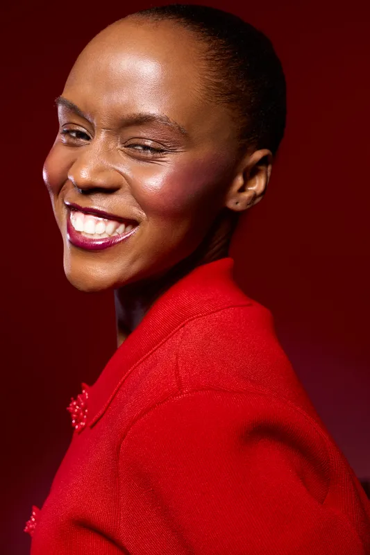 Smiling dark skinned African woman with short hair wearing red jacket winks and shows teeth on red background. Portrait of confident black female model expressing joy and positivity.