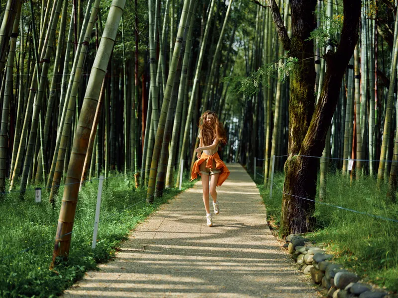 Woman running along a gravel path through a dense bamboo forest wearing a flowing orange dress, sunlight filtering between tall trees, motion and carefree summer nature scene.