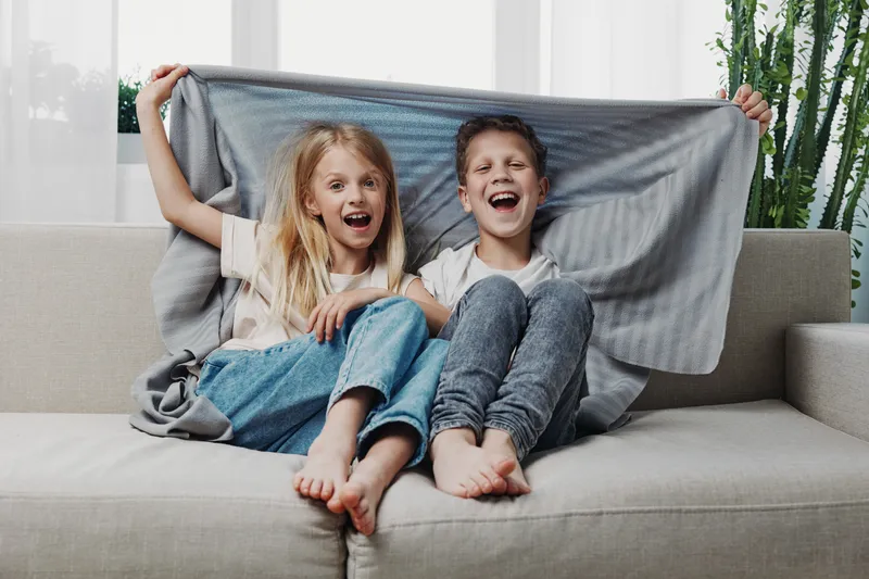Happy children sit barefoot on sofa covered with blanket, laughing joyfully in bright living room. Concept of childhood fun, friendship, and cozy home atmosphere with natural light.