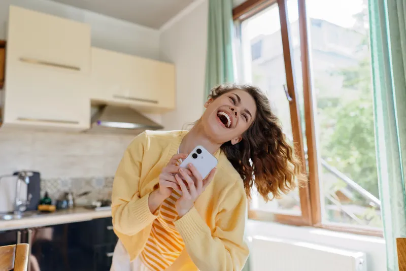 Cheerful woman in a sunny kitchen enjoys a moment at home, holding a smartphone and laughing, warm natural light creates a friendly, casual lifestyle portrait for domestic settings.