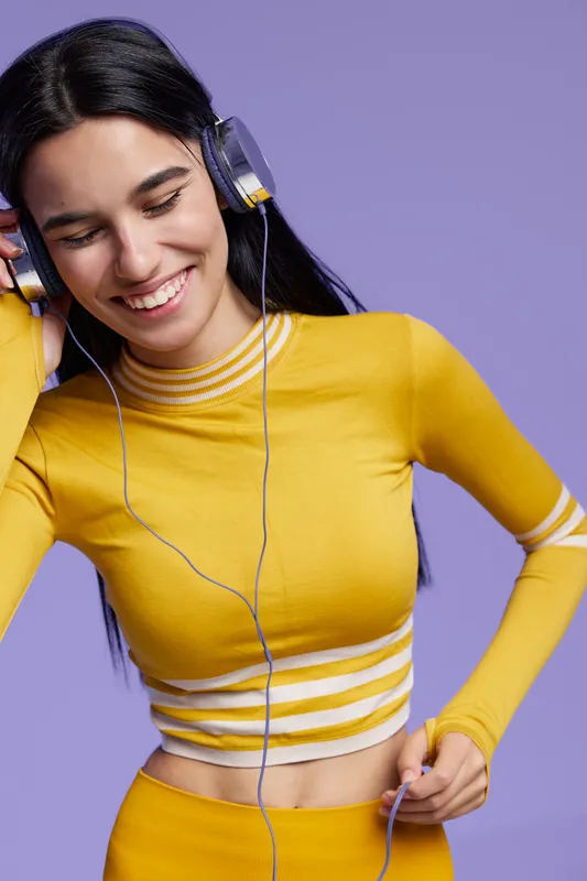 Young woman sport fitness enthusiast smiling while listening to music in yellow workout attire against a violet background, showing energy and active lifestyle.