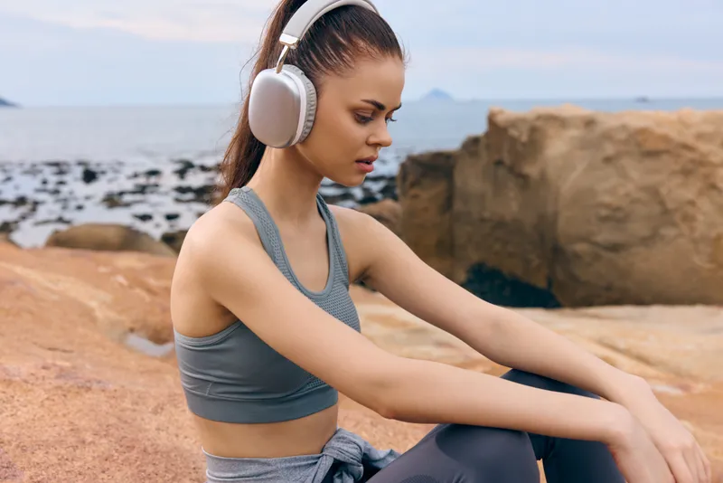 Young woman enjoying music with headphones by the beach, wearing sportswear, showcasing relaxation and serenity against a natural rocky backdrop