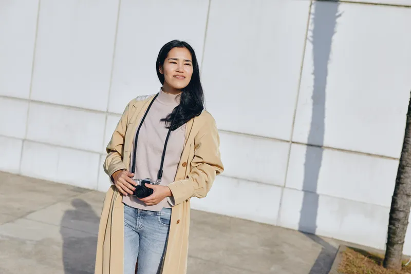 Woman with camera in a casual coat stands outdoors by a white wall, capturing a stylish street moment with natural light and confident stance. In the bright outdoor setting.