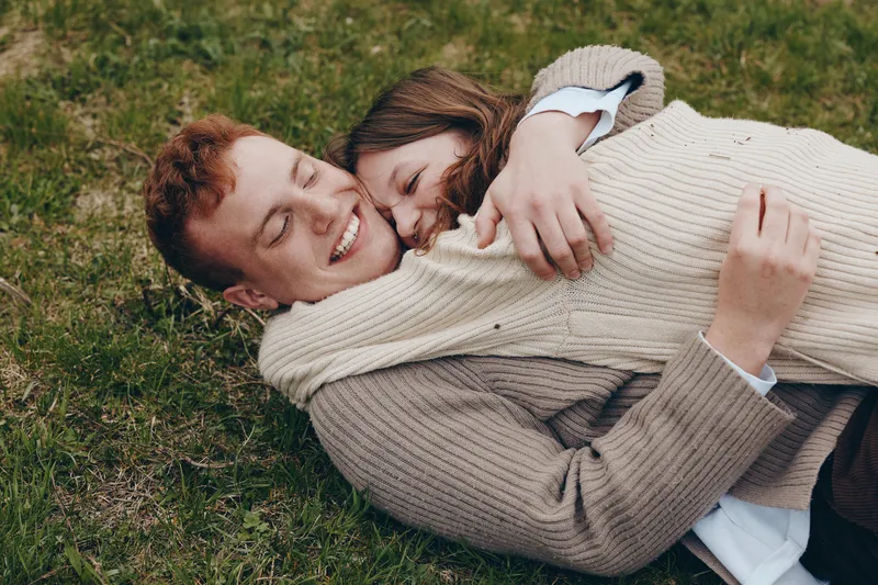 Two people lie on grass, sharing a warm hug and smiles, wearing knit sweaters, enjoying a cozy outdoor moment, natural light, candid lifestyle scene, joyful connection