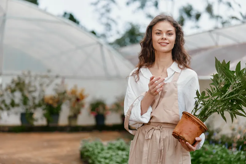 Young woman with curly hair wearing a white shirt and apron, holding a potted plant in a greenhouse, embodying a love for gardening and nature