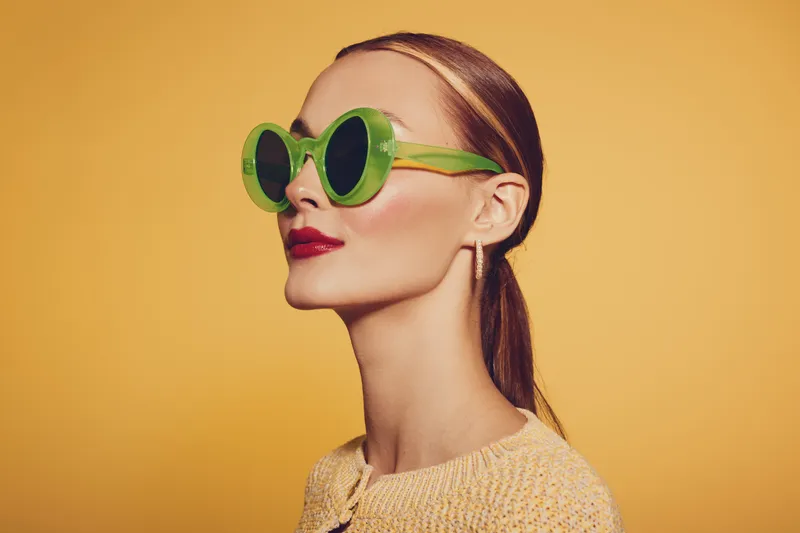 Stylish woman with green round sunglasses and red lipstick poses on yellow background. Fashion portrait with bright colors and modern accessories. Concept of summer style and beauty.