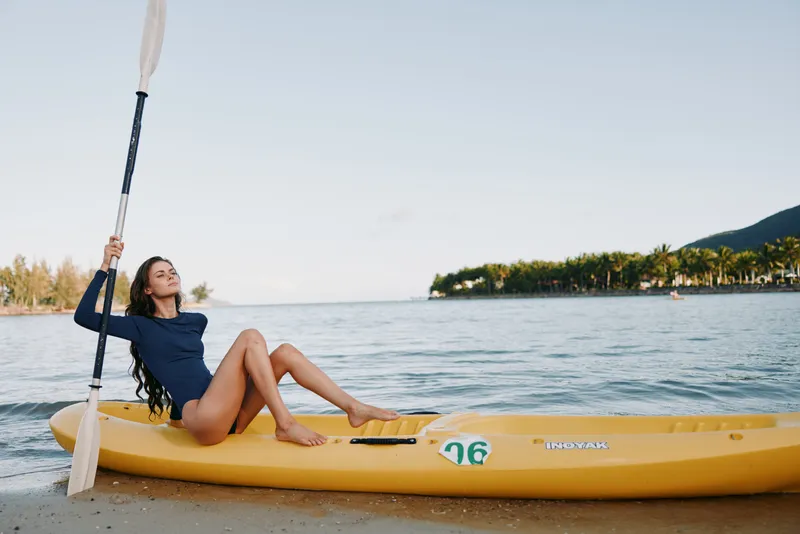 A young woman relaxes on a yellow kayak near the shore, paddle raised, calm water and clear sky creating a peaceful beach moment.