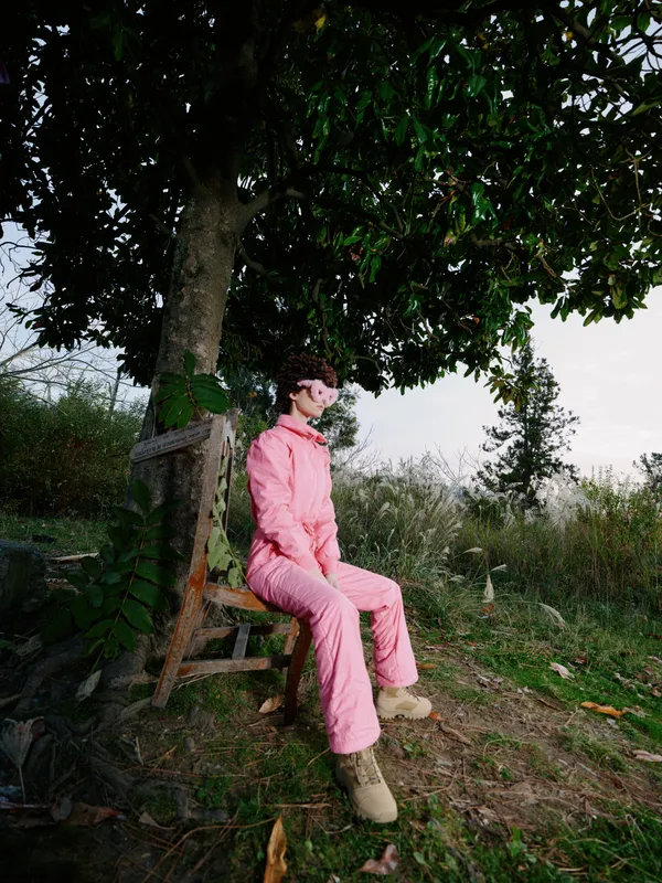 Outdoors scene with a person in a pink jumpsuit seated on a wooden bench under a leafy tree, relaxed pose in a natural garden setting with soft sunlight and greenery