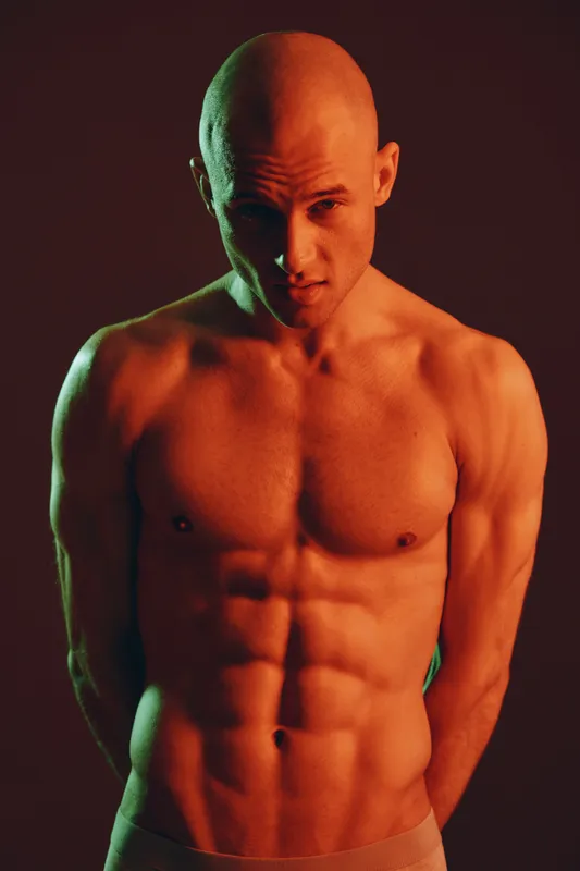 Muscular man posing dramatically in low light, showcasing impressive physique and intense expression. The image emphasizes strength and determination.