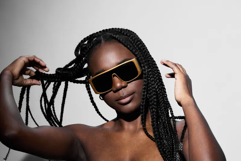 Confident young Black woman with braided hair and oversized sunglasses, posing against a minimalistic white background, exuding fashion forward style and individuality