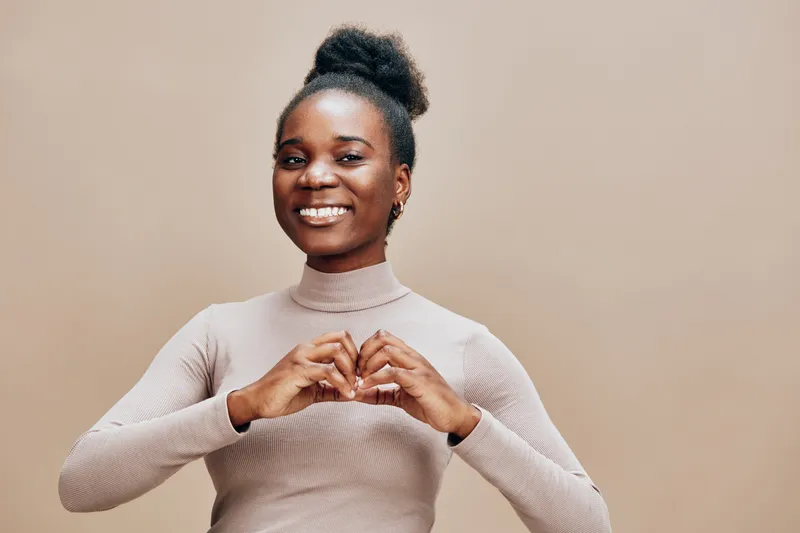 Smiling black woman making a heart shape with her hands against a neutral background, showcasing joy, love, and positivity in a stylish outfit