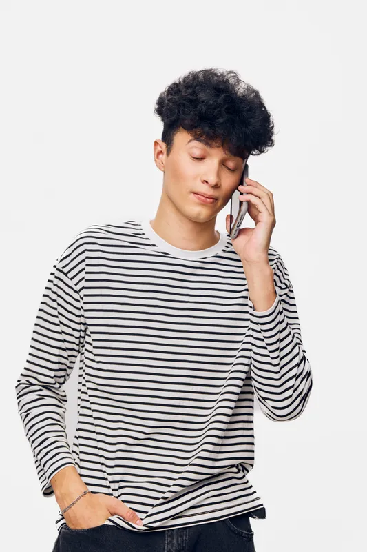 young man talking on smartphone wearing striped long sleeve shirt, casual style, curly hair, relaxed expression, light background, communication concept