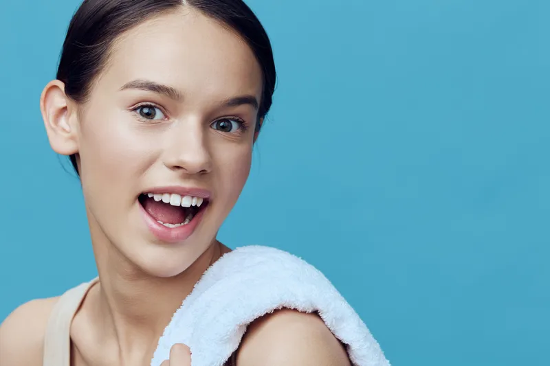Cheerful young woman with a towel on her shoulder, showcasing clear skin and bright smile against a blue background, embodying wellness and self care