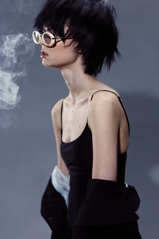Stylish woman with short hair wearing sunglasses and a black top, exhaling smoke against a dark gray background.