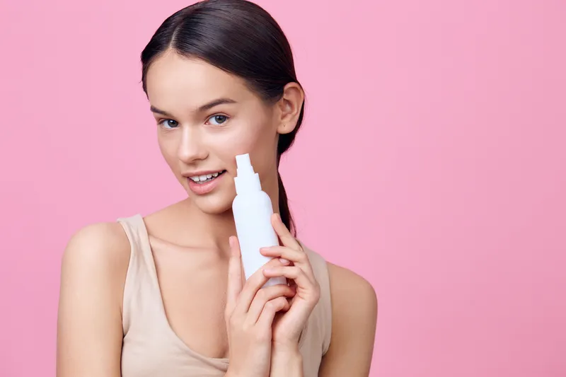 Young woman holding a skincare product against a pink background, showcasing her natural beauty and fresh skin Perfect for beauty and wellness themes