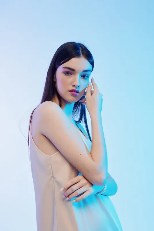 Young woman with long dark hair poses in a beige dress against a blue background. Soft lighting highlights her beauty and fashion style. Concept of elegance and modern beauty.