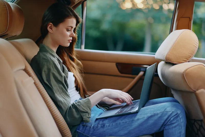 Young woman working on a laptop inside a car during sunset, portraying concentration and productivity in a chic casual outfit.