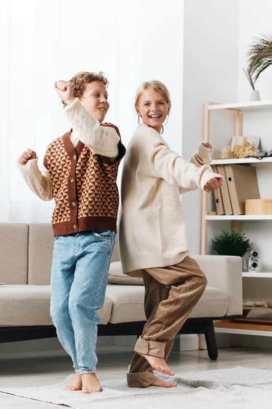 Two children dance happily indoors, wearing cozy sweaters and jeans, captured in bright natural light with a warm living room backdrop and a candid joyful vibe.