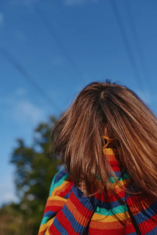 Child in colorful striped sweater hides face behind long hair in outdoor portrait, playful moment on a sunny day with blue sky and natural background.