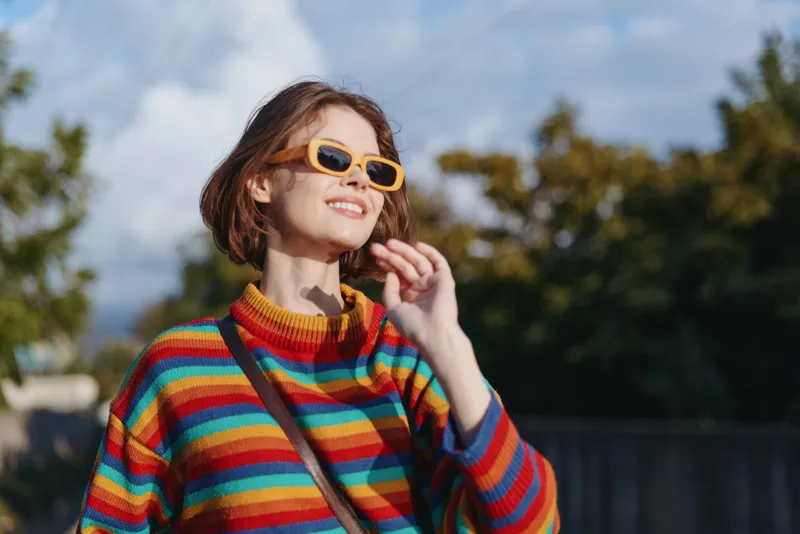 Woman smiling in a colorful striped sweater and yellow sunglasses, short hair and casual outfit walking outdoors in sunny suburban park, joyful travel lifestyle and fashion portrait.