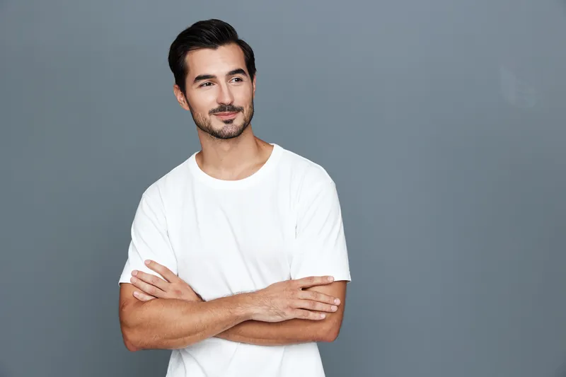 Smiling man with crossed arms looking confidently at the camera, dressed in a casual white t-shirt, captured against a soft gray background. Lifestyle concept.