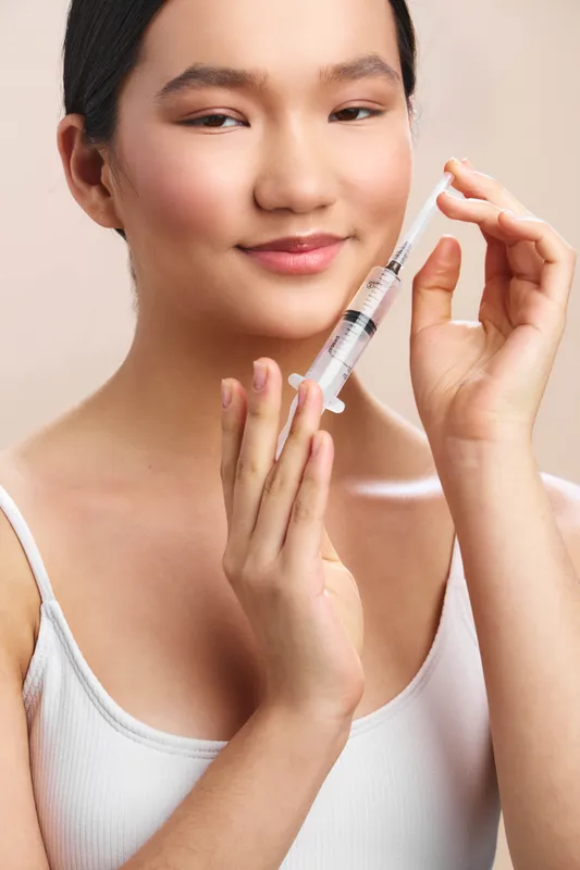 asian woman smiles softly as she holds a syringe near her face in a clean clinical setting, presenting confidence and beauty care in a modern wellness environment today properly