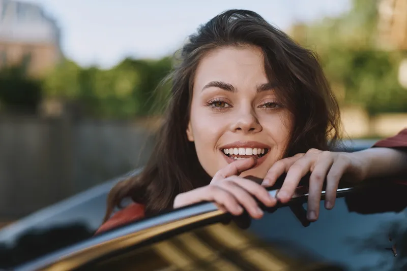 Smiling woman with brown hair leaning from car window, enjoying a sunny outdoor moment. Casual lifestyle scene with a playful mood, capturing freedom, spontaneity, and everyday happiness