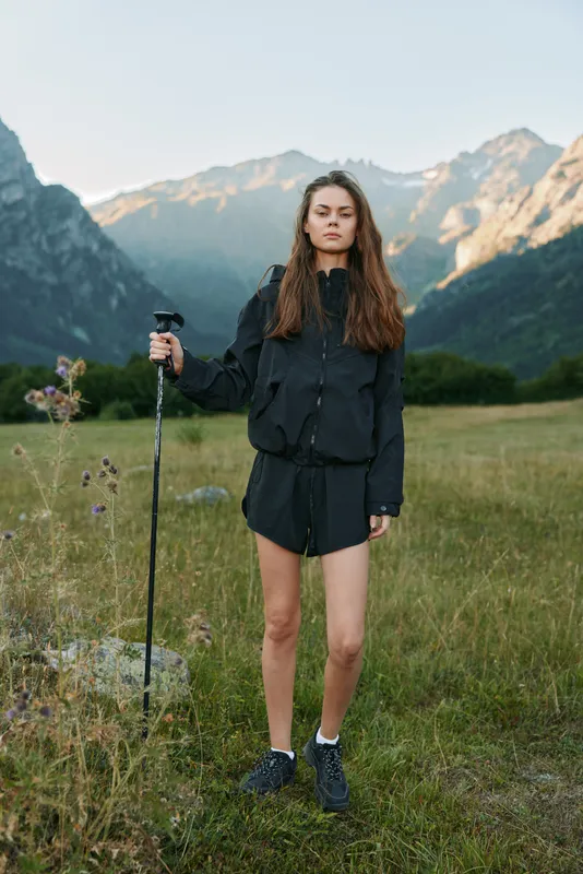 A lone young hiker stands in a mountain meadow, posing with a trekking pole. She wears a dark jacket and shorts, ready for adventure in rugged alpine scenery.