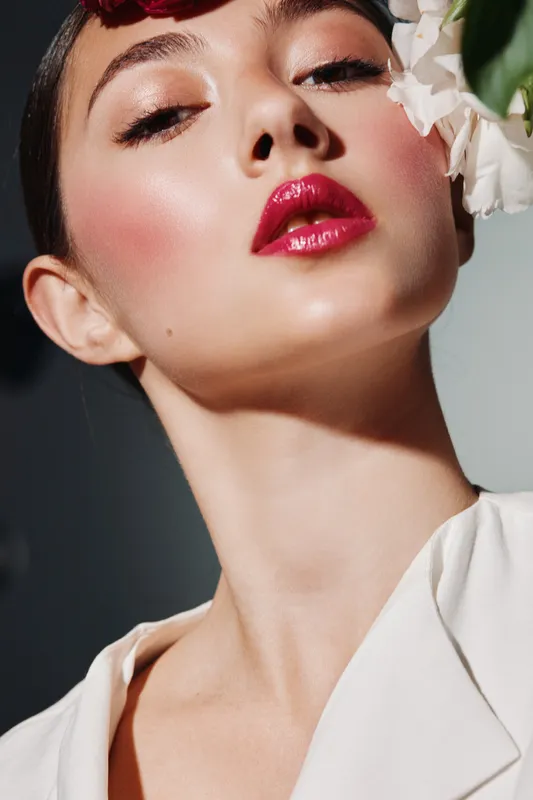 Young woman with glossy red lips and radiant skin posing in close-up fashion portrait. Bright makeup and artistic lighting highlight beauty and elegance. Creative beauty concept.