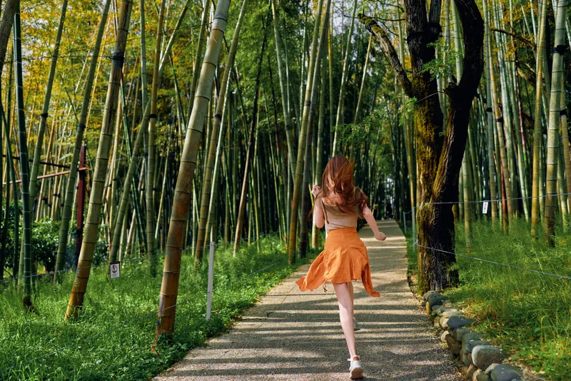 Woman running on a bamboo path wearing orange skirt, back view in nature forest. Summer sunlight filters through tall bamboo trees along a peaceful gravel trail, casual motion.