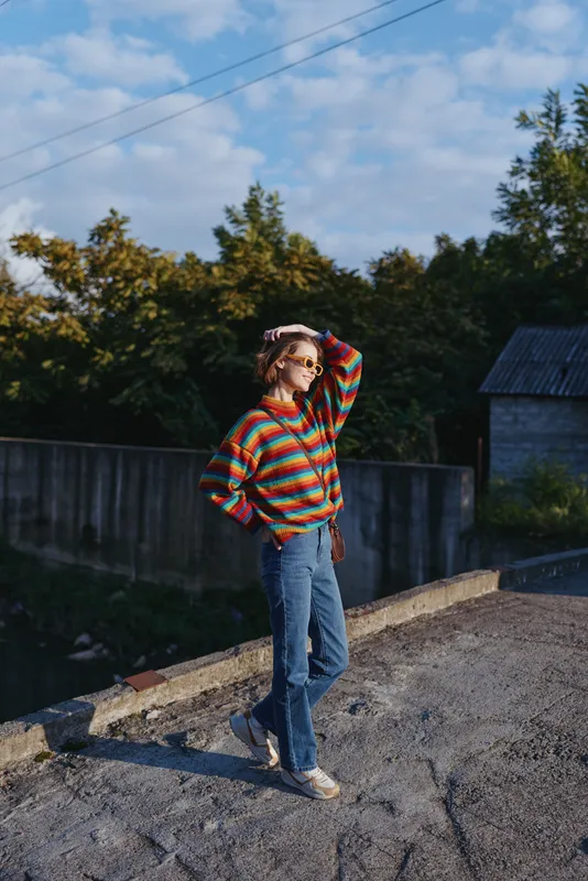 young woman in striped sweater and denim jeans wearing sunglasses, casual outfit, standing outdoors on roadside, early 20s, smiling and shielding eyes, relaxed travel lifestyle fashion vibe