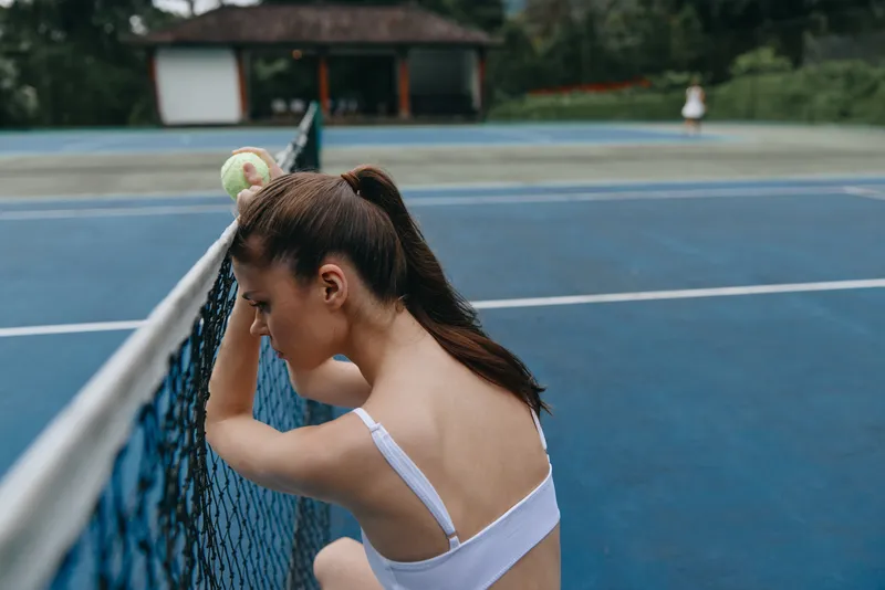 Young female athlete resting on a tennis court, wearing a white sports top, showing determination and focus while holding a tennis ball