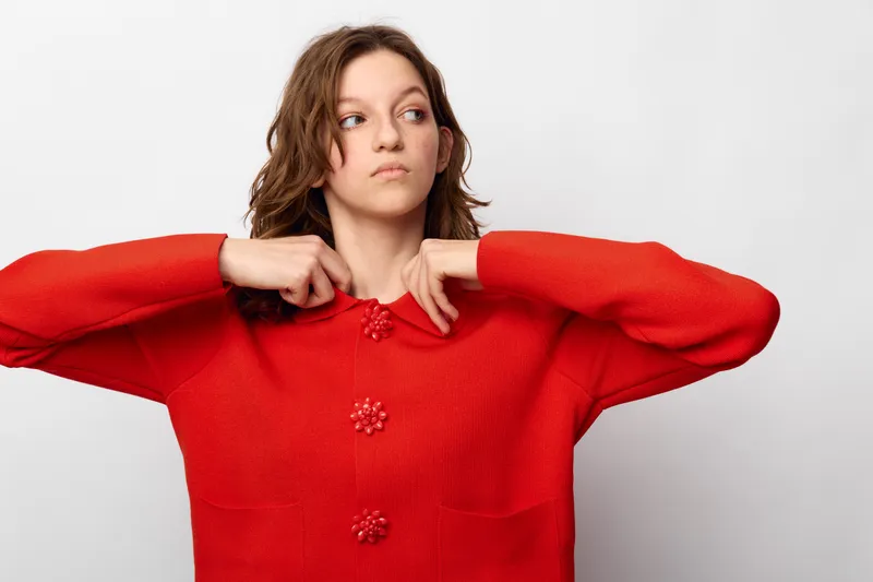 Young woman in red sweater adjusts collar looking sideways with thoughtful expression. Studio portrait on white background. Concept of fashion, casual style, and contemplation.