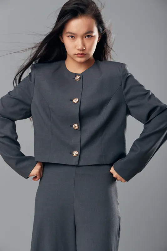 Woman posing in gray stylish suit with gold buttons looking confidently at camera with serious expression, long dark hair and hands on waist in studio setting.
