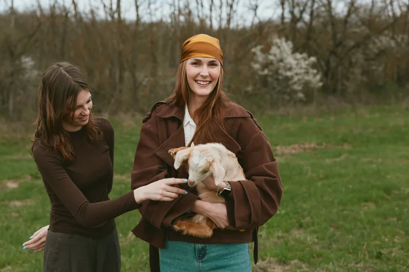 Two women outdoors in a park share a moment as one holds a small dog, both in casual autumn outfits, smiling warmly, creating a lively lifestyle scene of friendship and pet companionship.