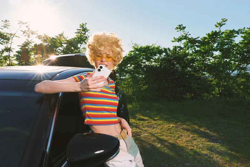 Cheerful woman outdoors with striped top leaning on a car, capturing a moment with a compact camera, sunlit scene, colorful retro vibes, casual lifestyle, soft breeze