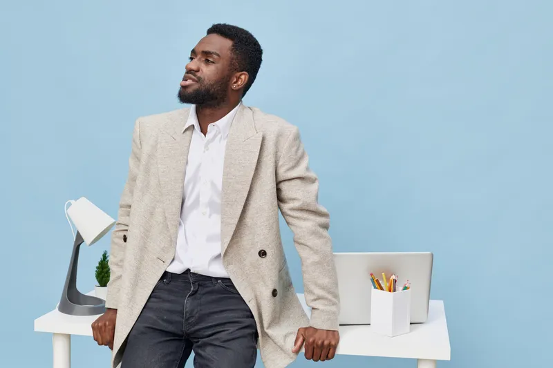 Confident man in smart casual outfit poses relaxedly at home office workspace with modern computer, white desk, and stationery, against pastel blue background. Lifestyle, professional, happiness conce