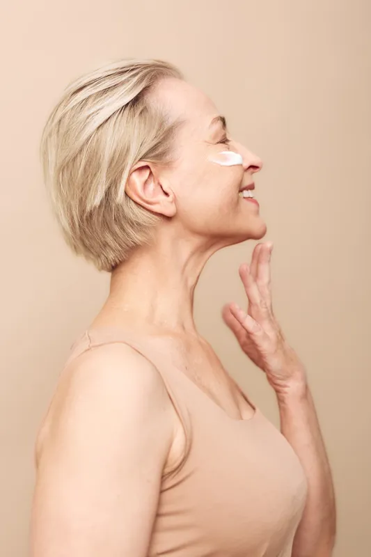 Profile of happy mature woman with short blonde hair applying white face cream on cheek. Soft beige background and natural makeup emphasize skincare, beauty, and self-care concept.