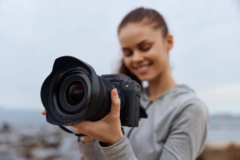 Young female photographer smiling while holding a camera by the seaside, showcasing her passion for photography, nature, and creativity in a casual outfit