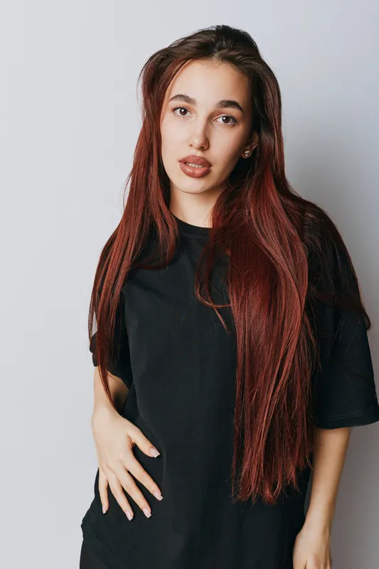 Young woman with long red hair wearing black t shirt standing against white background looking at camera with neutral expression and hand on hip in studio portrait.