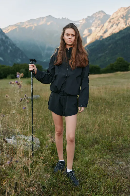 A focused young woman stands in a scenic alpine meadow with mountains behind. She holds a trekking pole, dressed in dark athletic wear, ready for hiking and exploration.