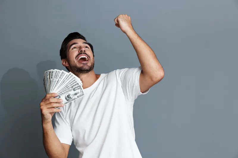 Happy man celebrating success while holding stack of dollar bills in hand. His joyful expression radiates excitement and achievement in a minimalist gray background.
