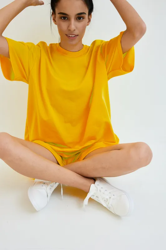 young woman in bright yellow oversized t-shirt sitting cross-legged on white floor with casual white sneakers, relaxed pose and natural expression indoors