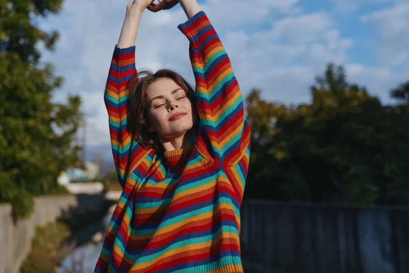 Woman in rainbow sweater stretching outdoors with eyes closed, portrait of relaxed young adult enjoying sunny day, casual fashion and serene mood in nature, warm golden light