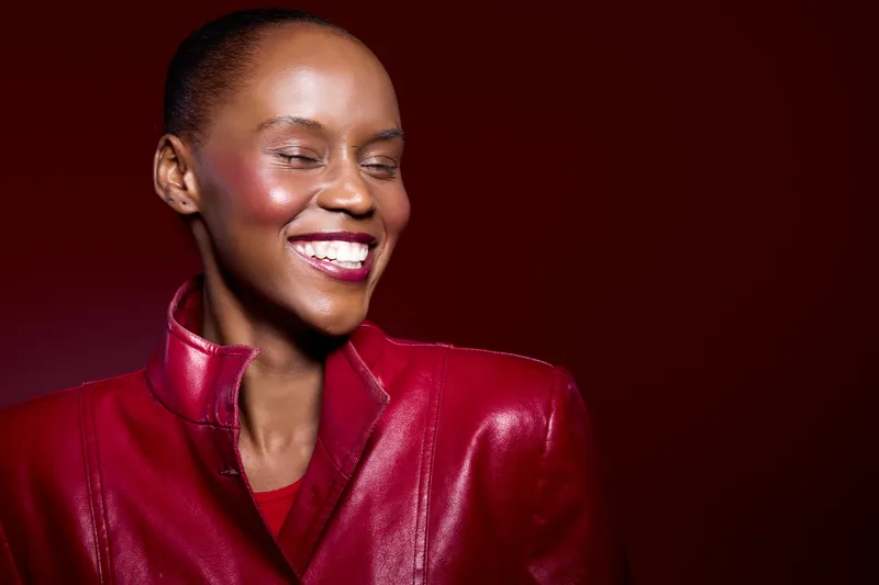 Smiling African woman with dark skin wearing red leather jacket on dark red background. Portrait with closed eyes and bright makeup. Concept of fashion, joy, and style for dark skin tone.