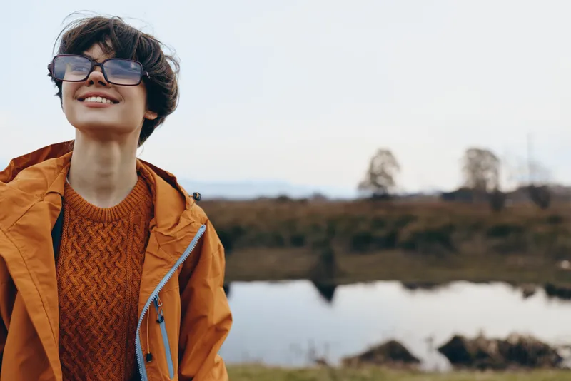 woman lifestyle outdoor smile with glasses and coat, standing by river in nature, enjoying daylight, relaxed gesture outdoors, soft wind, scenic landscape background, candid moment